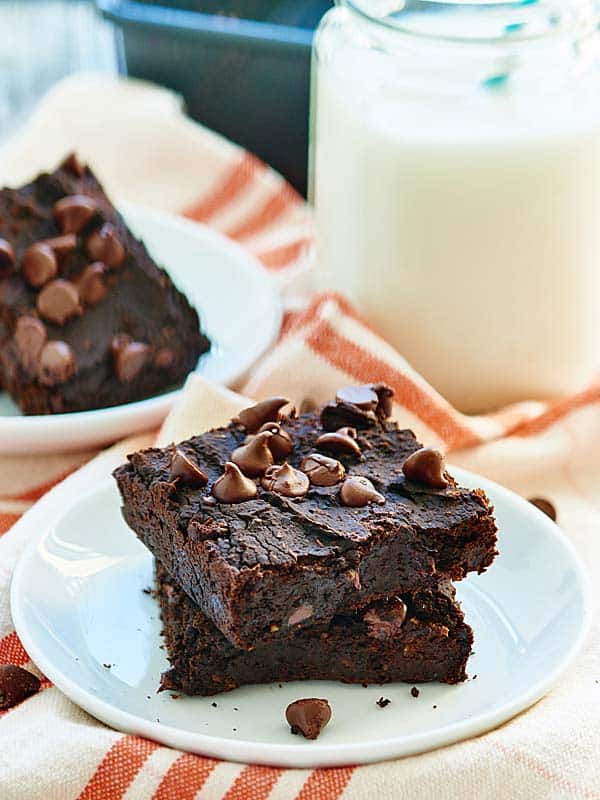 Two brownies stacked on a plate with milk and another brownie in the background