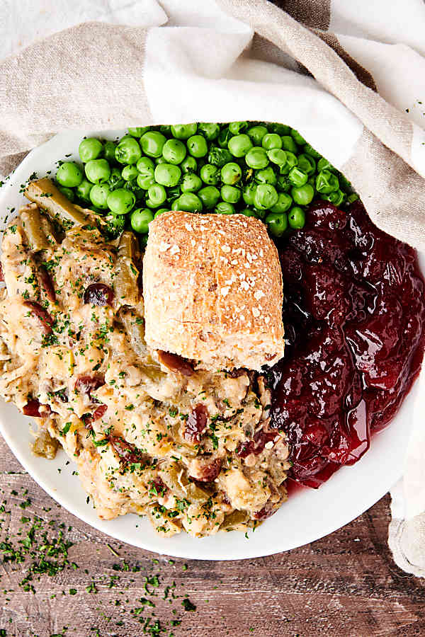 crockpot chicken and stuffing on plate with cranberry sauce, peas, and dinner roll above